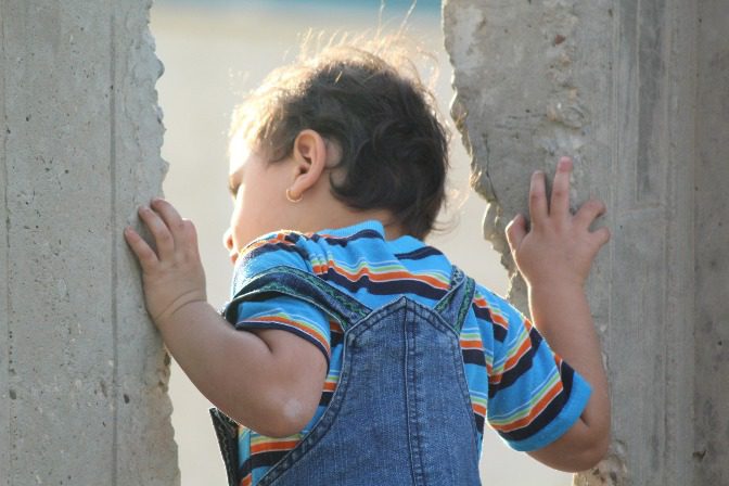 Child peering through concrete wall gap.